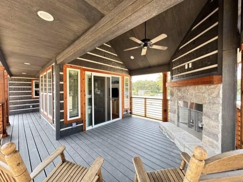 A rustic covered porch with wooden chairs, a ceiling fan, stone fireplace, and glass sliding door. The ambiance is cozy and inviting, overlooking greenery.