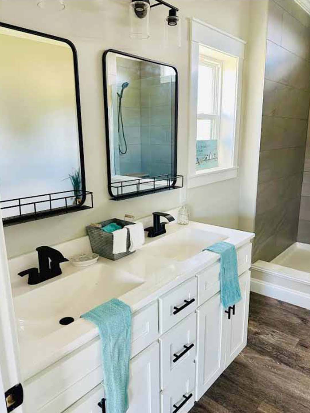 Modern bathroom with dual sinks and black faucets. Two mirrors above feature small shelves. White cabinetry contrasts with light teal towels and warm wood floor. Relaxing ambiance.