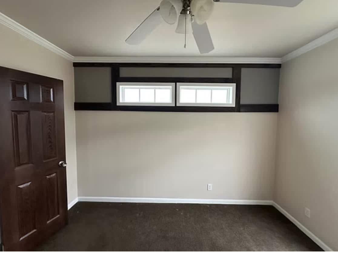 A small, empty room features white walls, a dark wooden door, and a brown carpet. Two narrow horizontal windows provide natural light, while a ceiling fan hangs above.