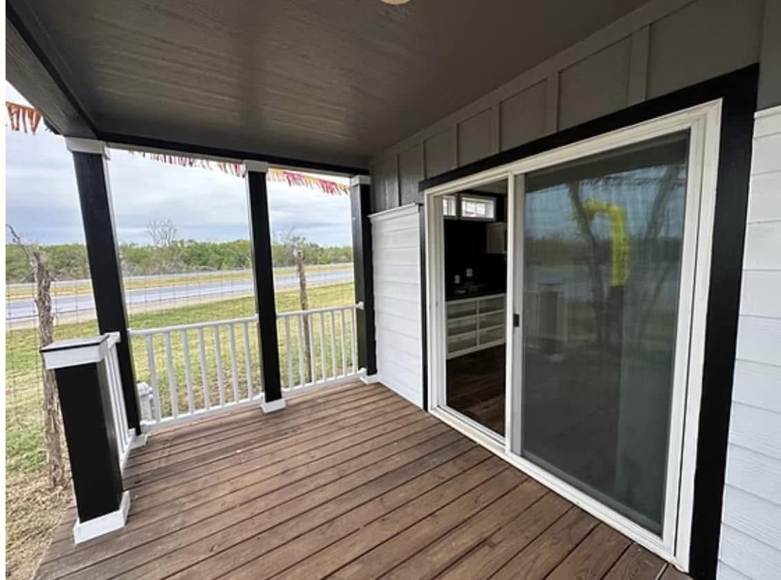 Cozy covered porch with wooden floorboards and white railings. A sliding glass door opens to the interior. Peaceful, rural setting with greenery visible.