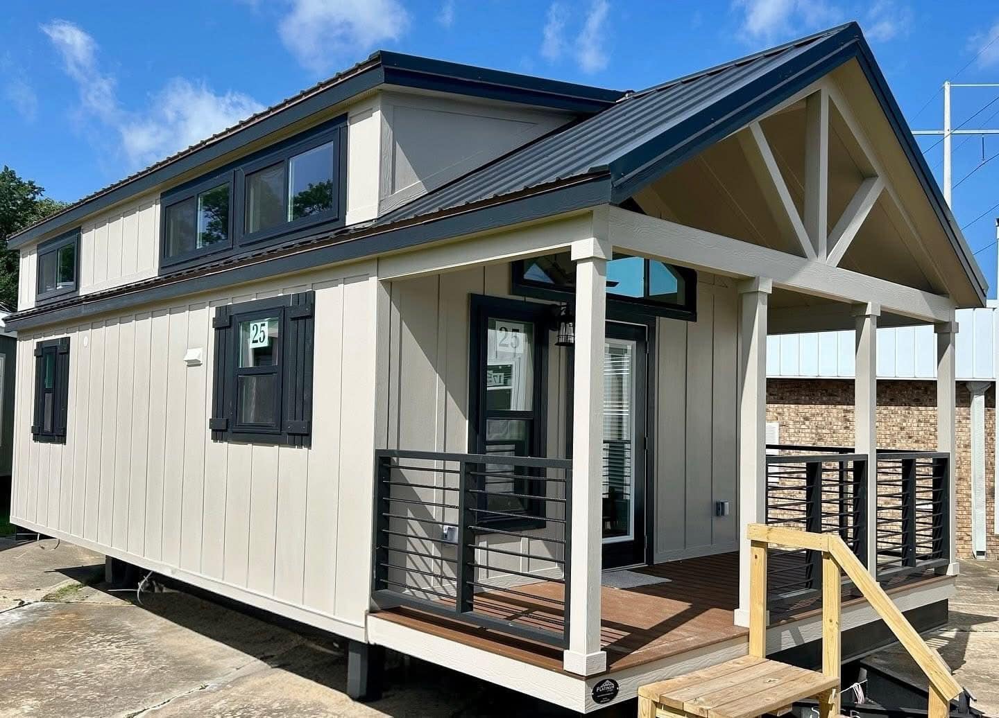 A modern tiny house with a gabled roof and beige siding. It features large windows, a small porch with black railings, and a wooden entry ramp.