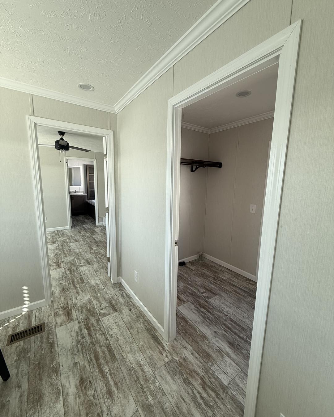 Hallway with light wood flooring and cream walls. A door to a closet on the right, and another room with a ceiling fan and bathroom visible further down. Calm, neutral tone.