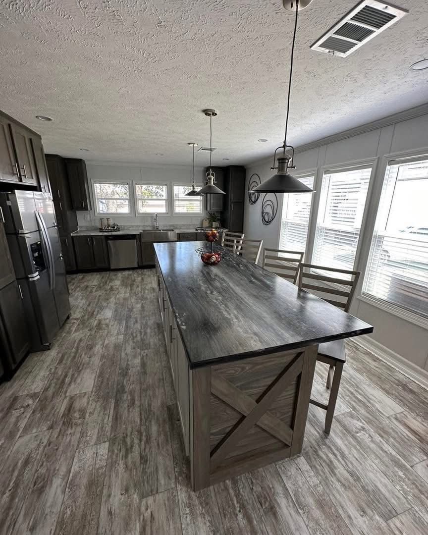 Modern kitchen with a large island featuring a dark countertop and wooden base. Pendant lights hang above. Bright windows line the right wall.