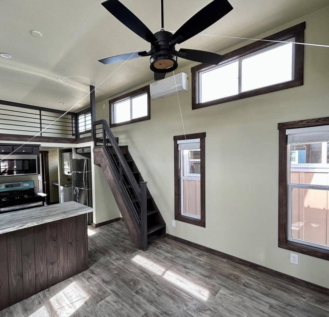 Modern tiny home interior with high ceilings, large windows, a ceiling fan, wood floors, a dark wood staircase to a loft, and a sleek kitchen.
