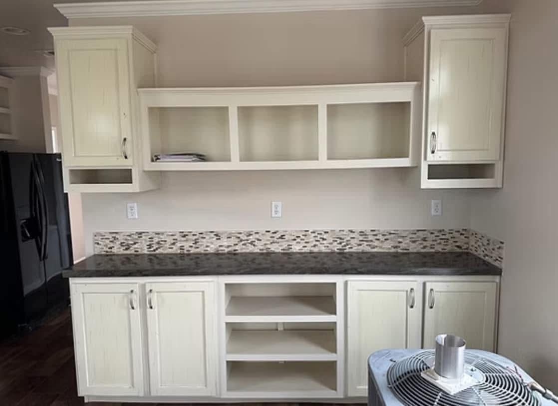 Cream-colored kitchen cabinets with black countertops, featuring open shelving and a mosaic tile backsplash. A hint of modern, cozy design.