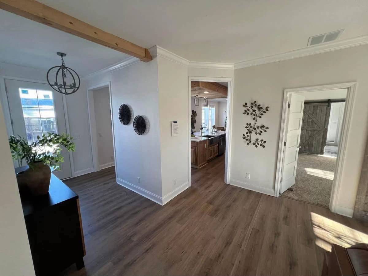 Hallway with wooden floor, white walls, and decorative wall art. A door leads to a modern kitchen and another to a cozy room with carpet. Bright, airy ambiance.