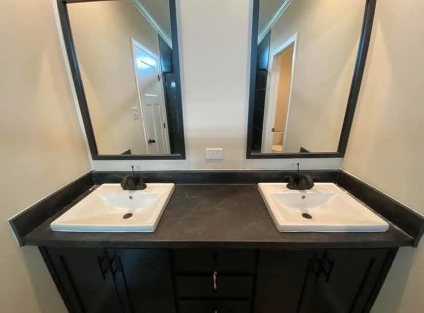 Modern bathroom vanity with dual white sinks and black faucets on a dark countertop. Two large rectangular mirrors reflect a softly lit hallway.