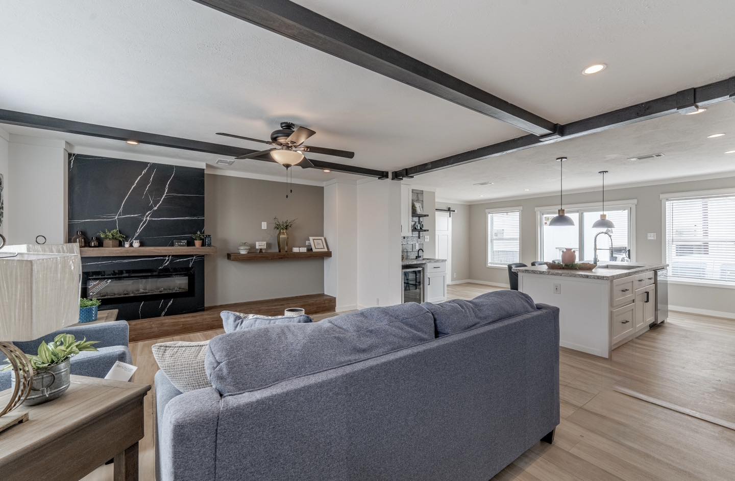 Spacious modern living room and kitchen with open layout. Gray sofa, sleek black fireplace, island with pendant lights, and large windows. Bright and inviting.