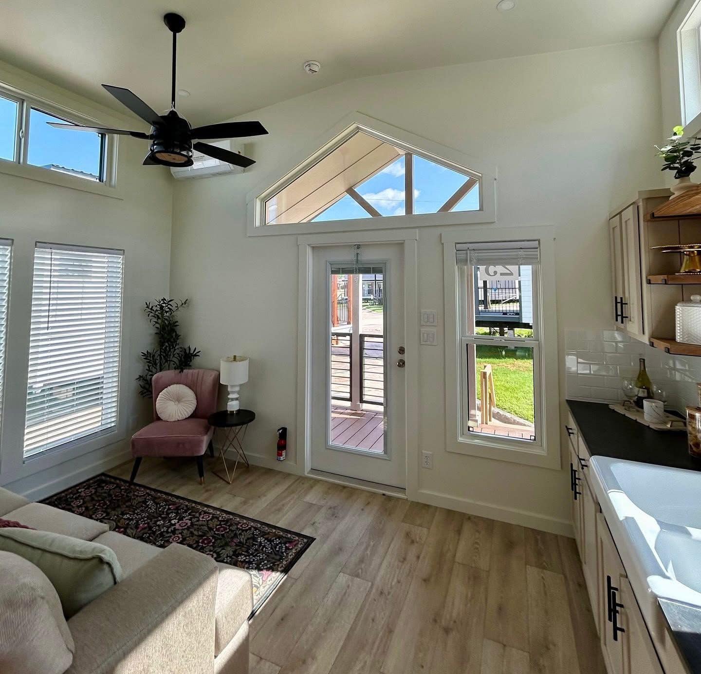 Bright living room with high ceilings, ceiling fan, and large windows. Includes a pink chair, stylish rug, and kitchen counter, exuding a cozy, modern vibe.