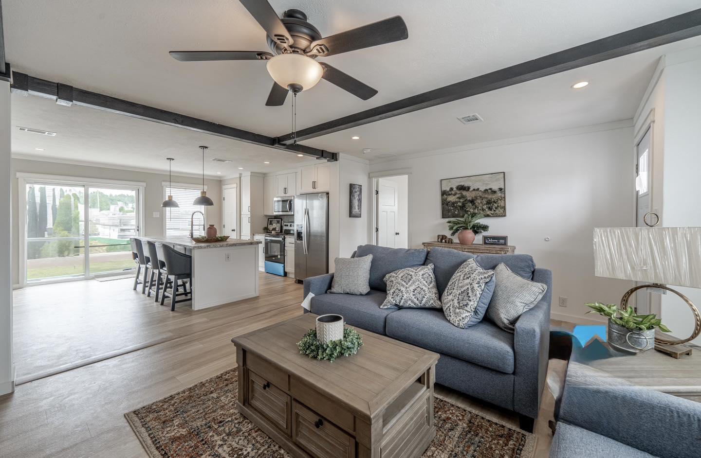 Modern living room with cozy navy sofa, decorative pillows, and wooden coffee table. Open layout leads to a bright kitchen with large windows.