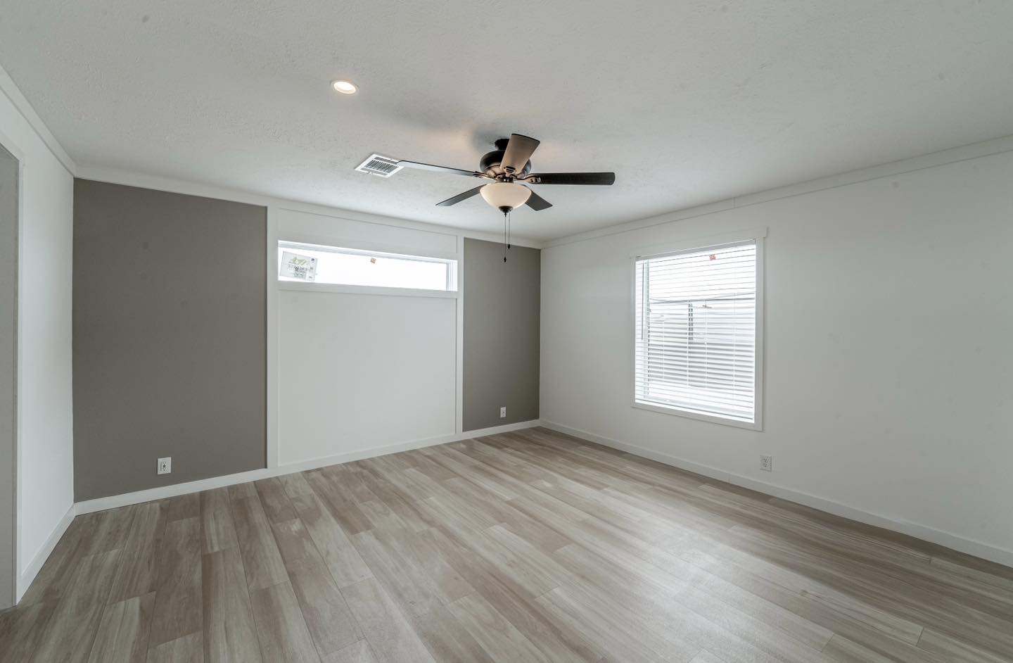 Spacious, empty room with light wood flooring, gray and white walls. A ceiling fan with lights is centered. Window blinds allow soft daylight in.