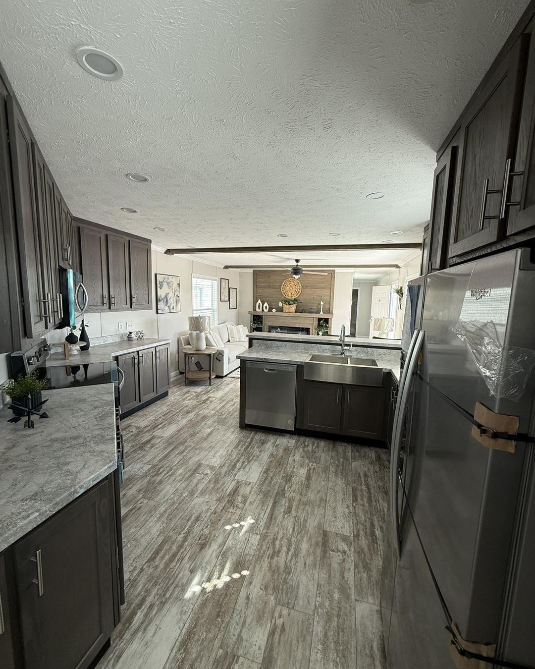 Modern kitchen and living area with wooden floors, dark cabinets, and stainless steel appliances. Bright natural light and cozy seating enhance warmth.