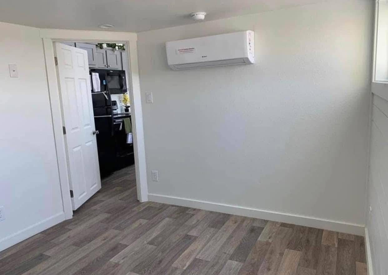 Empty room with gray wood flooring, a white wall-mounted air conditioner, and an open door revealing a small adjoining kitchen area. Bright and minimalist.