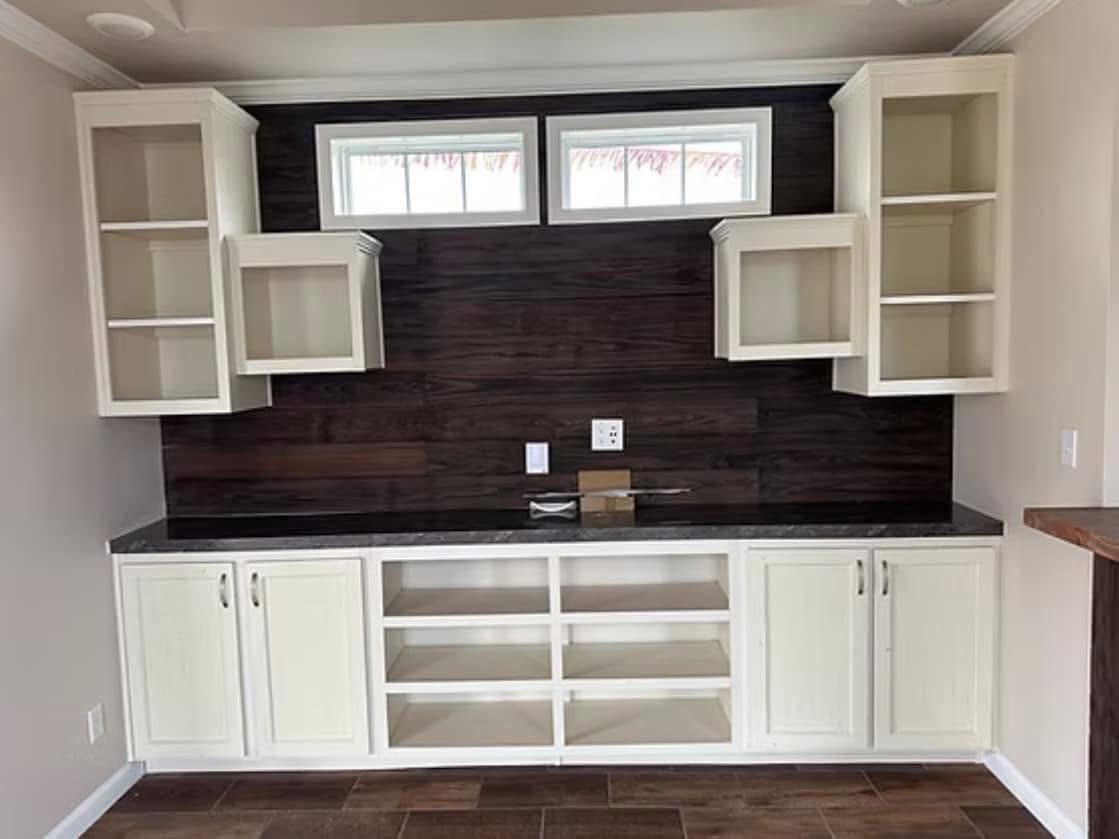Modern kitchen with open white cabinets, dark wooden backsplash, and light countertops. Three small windows above add natural light. Minimalist feel.