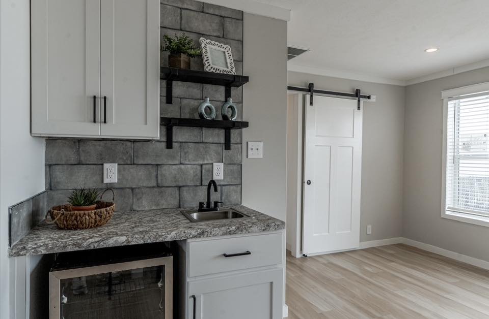 Compact kitchenette with marble countertop, small sink, and mini fridge. Gray tile backsplash, black shelves with decor. Sliding door and wood floor.