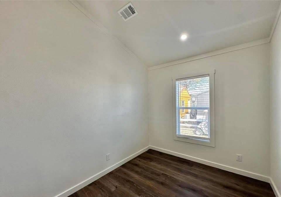 A small, empty room with white walls and dark wood floors. A single window lets in natural light, revealing a bright exterior and a cozy atmosphere.
