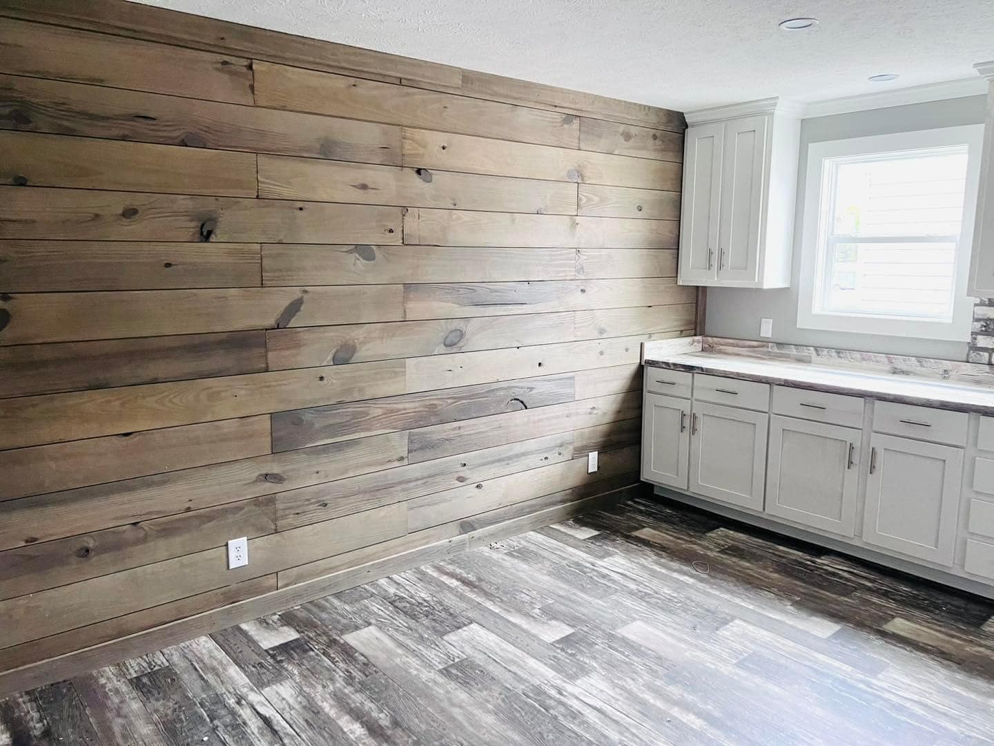 Empty room with rustic wooden wall paneling and gray cabinetry. A window above the counter lets in natural light, creating a cozy atmosphere.