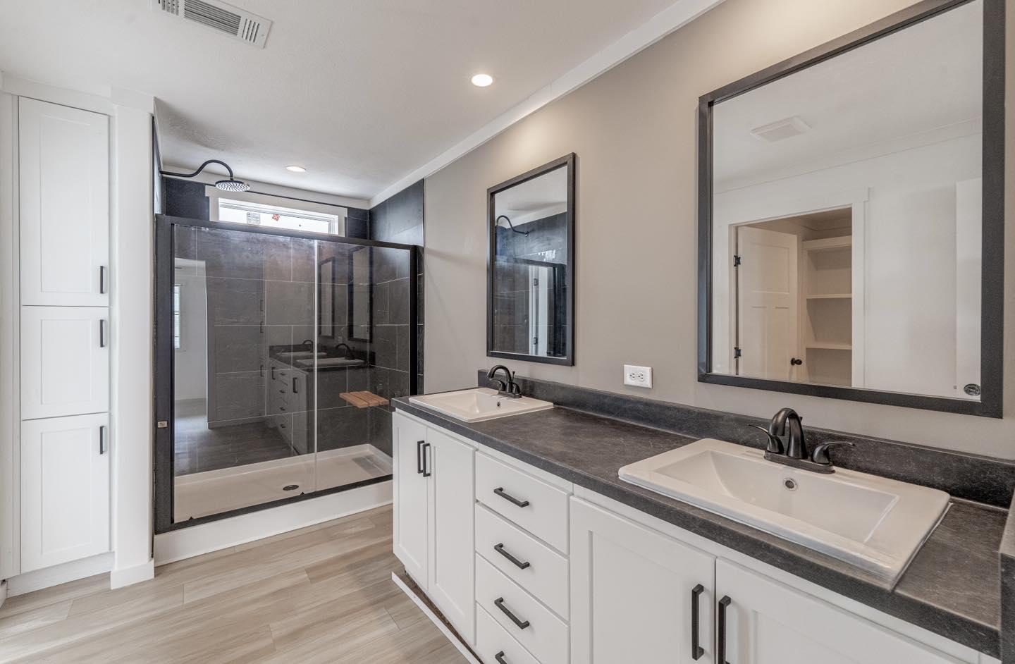 Modern bathroom with dual sinks on a sleek black countertop, a large mirror above each, and a glass-enclosed shower with dark tiles in the background.