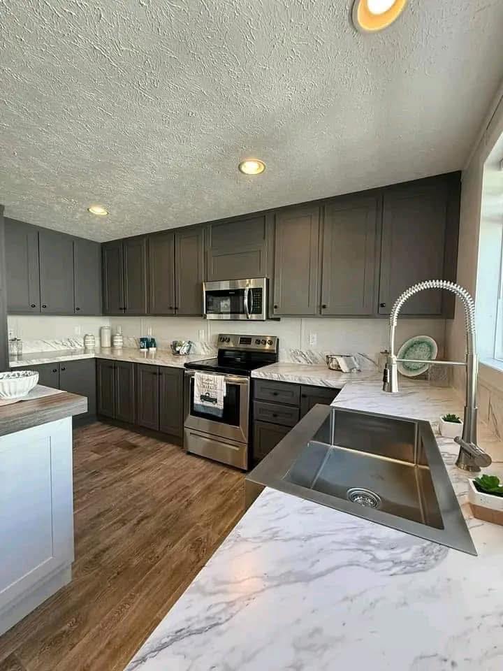 Modern kitchen with gray cabinets, stainless steel appliances, and marble countertops. Wood flooring adds warmth. Sleek faucet over a deep sink. Cozy and stylish.