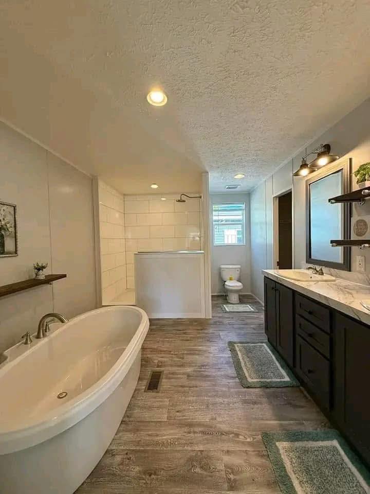 Spacious bathroom with a white freestanding tub, wooden floors, shower stall, and double-sink vanity. Warm lighting and green accents create a serene ambiance.