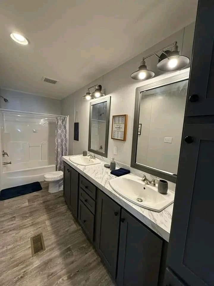 Modern bathroom with dual sinks on a marble countertop, two mirrors, and gray cabinets. Bathtub with shower and curtain. Warm, inviting lighting.