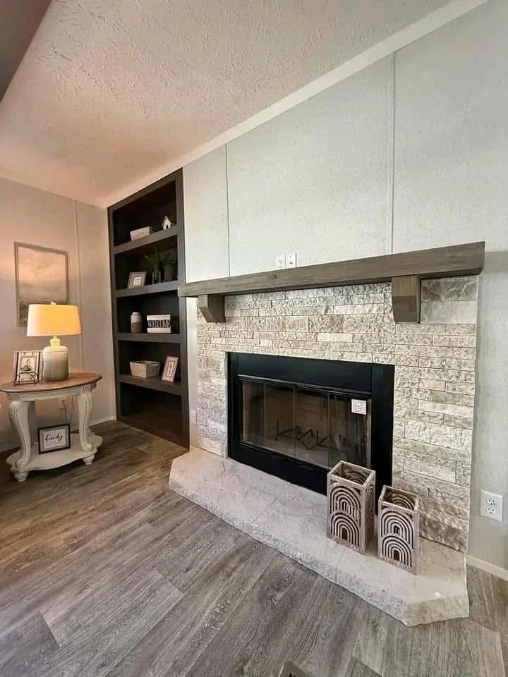 Cozy living room with a stone fireplace, wooden mantle, and decorative vases. A warm lamp on a side table and dark shelving create an inviting atmosphere.