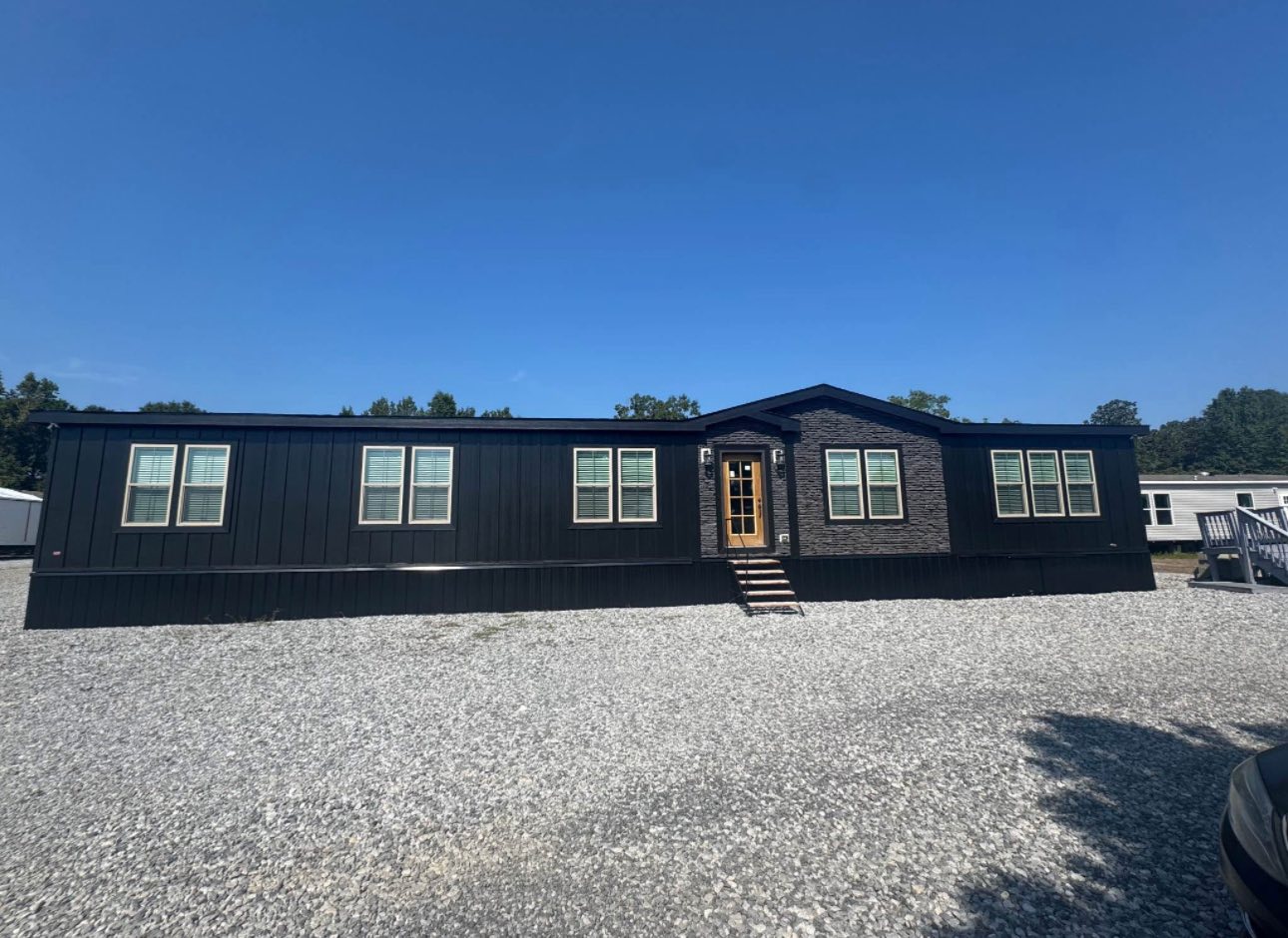 Single-story black modular home with brick accents, six windows, and a wooden door, set against a clear blue sky. Surrounded by gravel driveway.