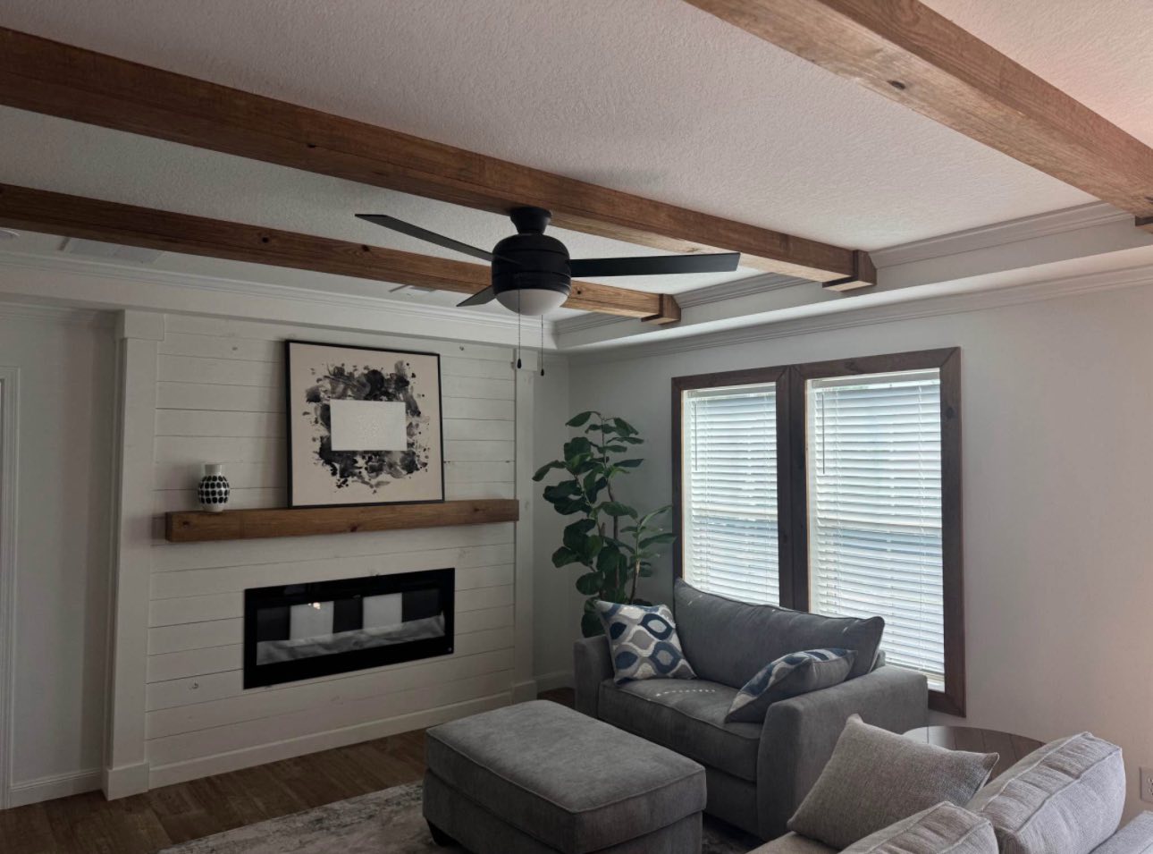 A cozy living room with a gray sectional sofa and patterned pillows. Wooden beams and a ceiling fan add warmth. Sunlight filters through large windows.