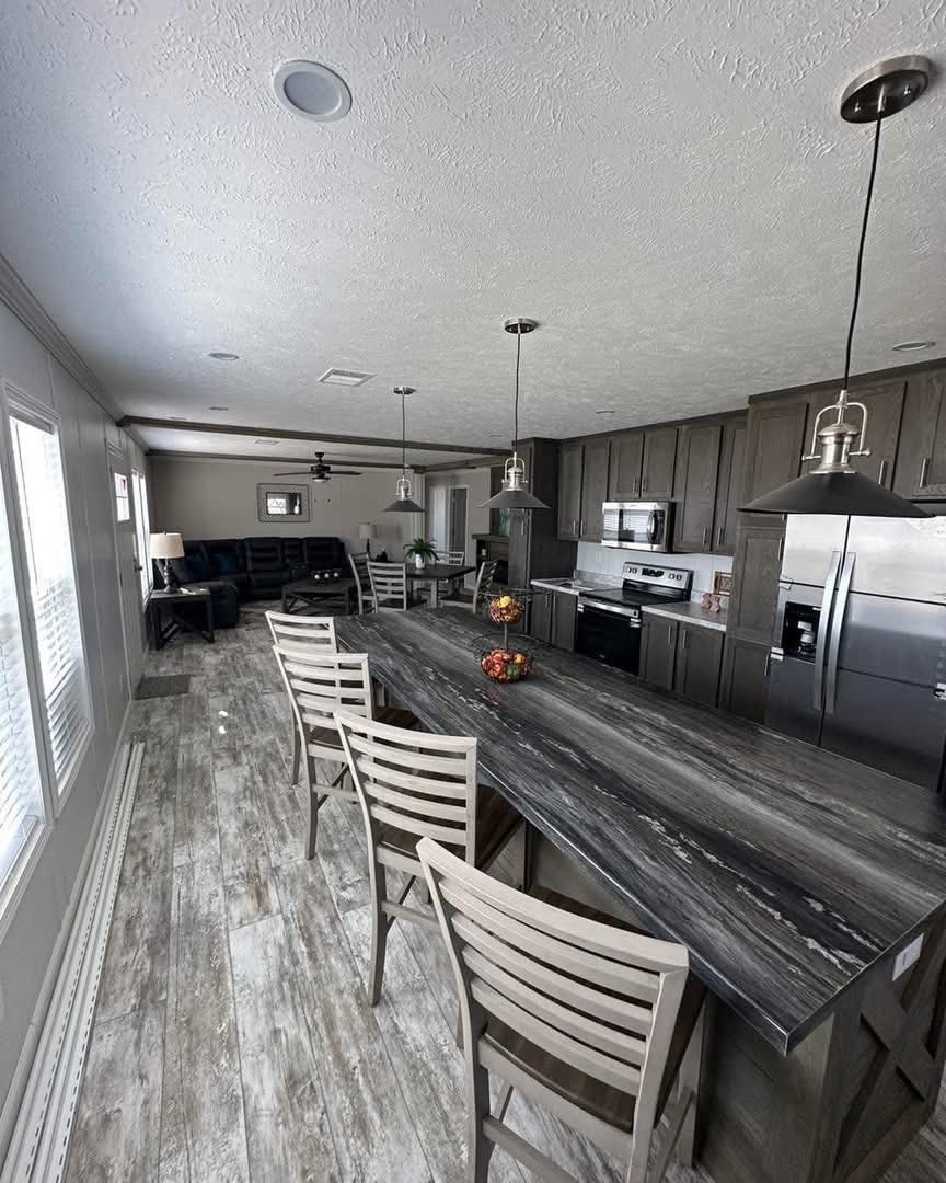 Spacious kitchen-living area with dark wood cabinets, stainless steel appliances, gray island, and rustic flooring. Cozy seating and natural light create warmth.