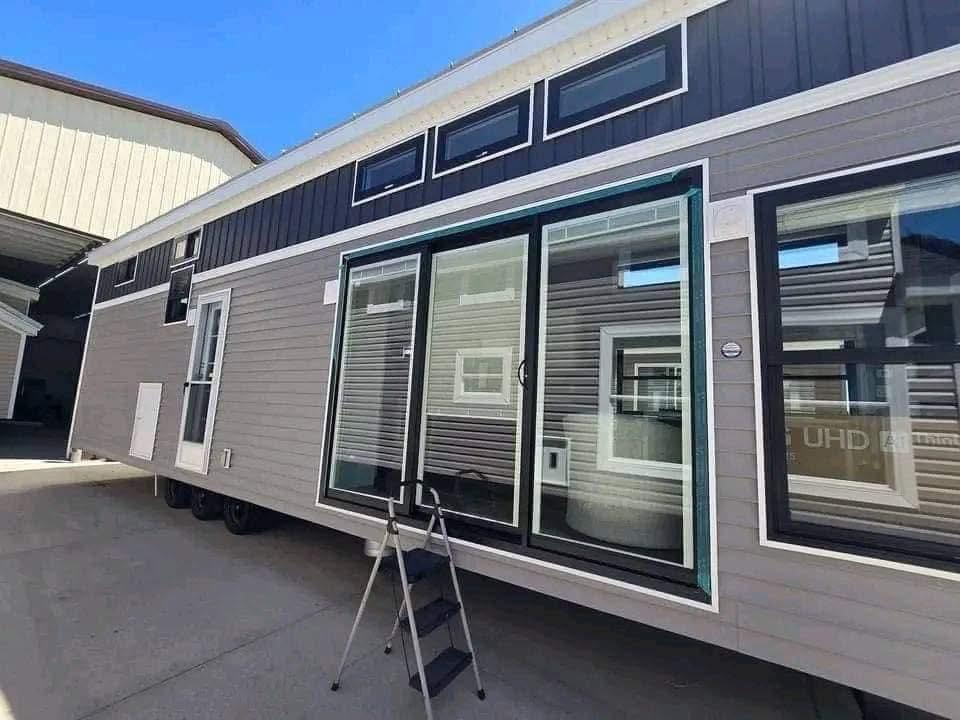 Modern tiny house exterior with gray siding and large glass sliding doors. Upper small windows provide light. A portable step ladder sits in front. Bright and sunny day.