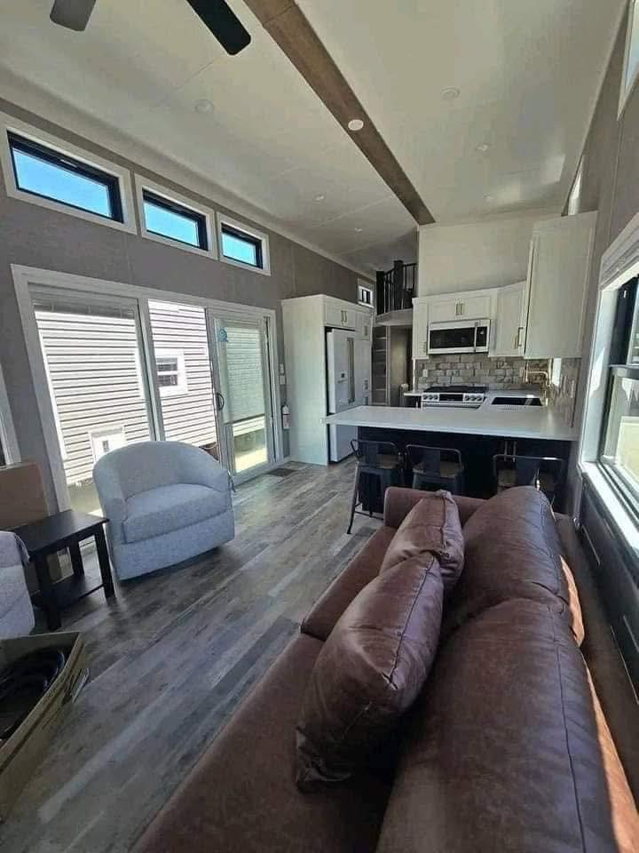 Modern tiny home interior with vaulted ceiling, featuring a brown sofa and gray chair, compact kitchen with white cabinets, and bright natural light.