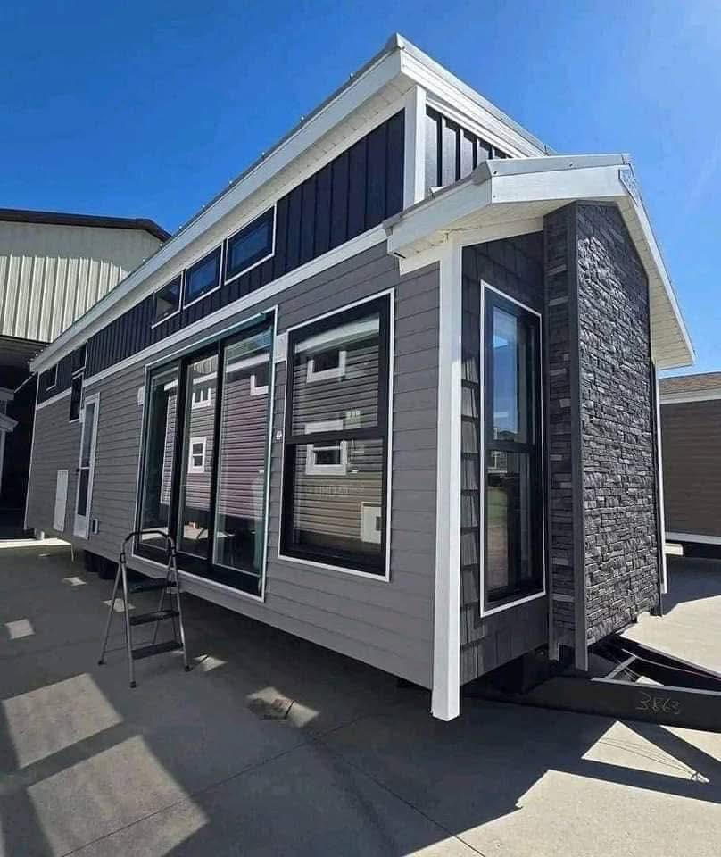 Modern tiny house with large windows and gray siding under a clear blue sky. A stone facade and a small ladder are visible, conveying a clean, minimalist look.