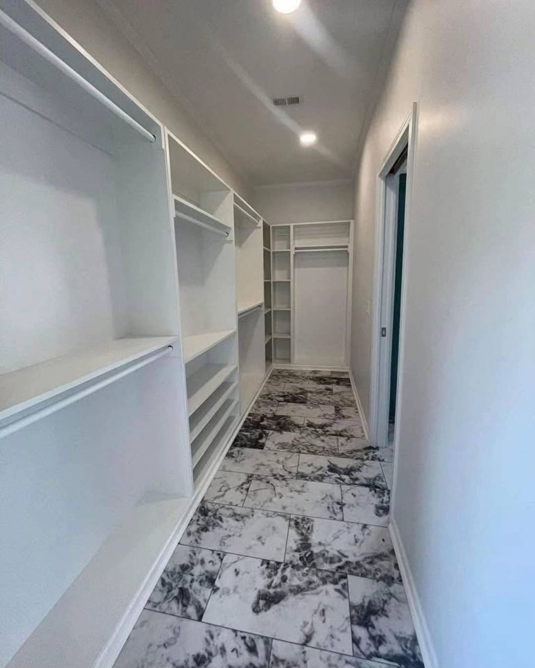 Narrow walk-in closet featuring white shelving and cabinets on the left and marble-patterned tile flooring. The space appears bright and orderly.
