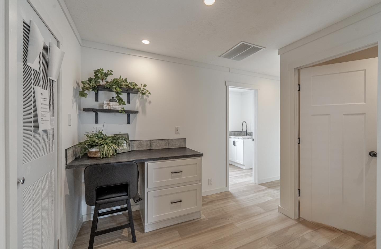 Minimalist home workspace with a small desk and black chair, flanked by drawers. Two shelves with green plants adorn the wall. Bright and organized.