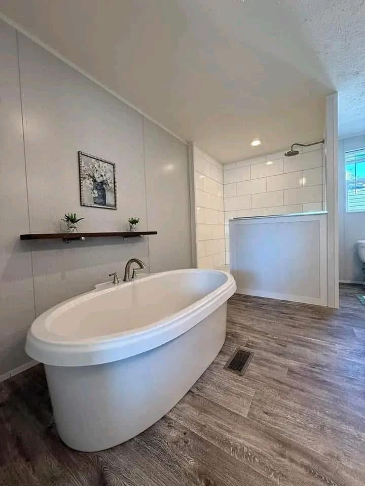 Modern bathroom with a freestanding oval bathtub on wood flooring. A shelf with potted plants and art decorates the wall. Shower and toilet visible.