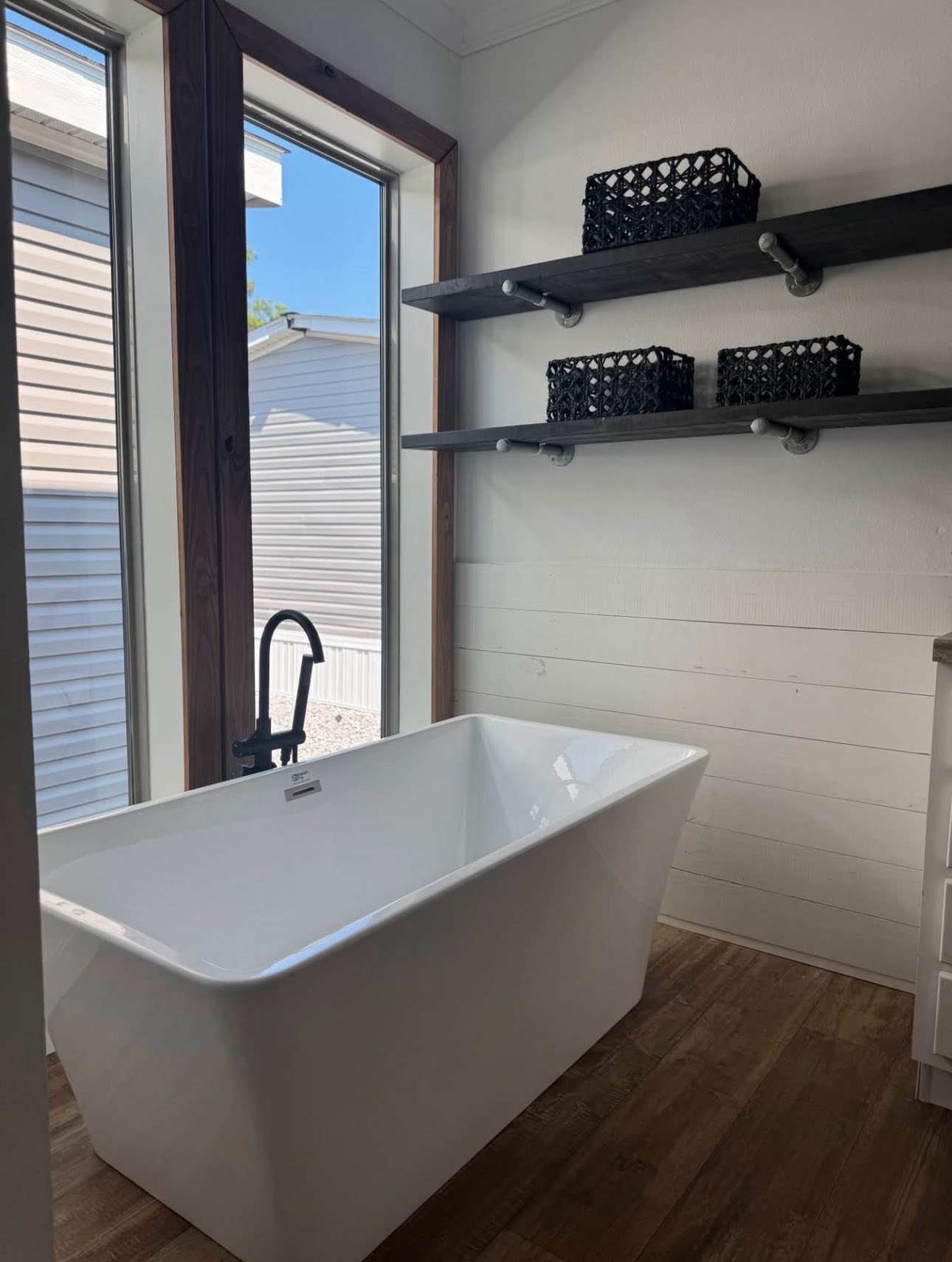 A modern bathroom features a white rectangular bathtub with a black faucet, large windows allowing natural light, and black woven baskets on wooden shelves.