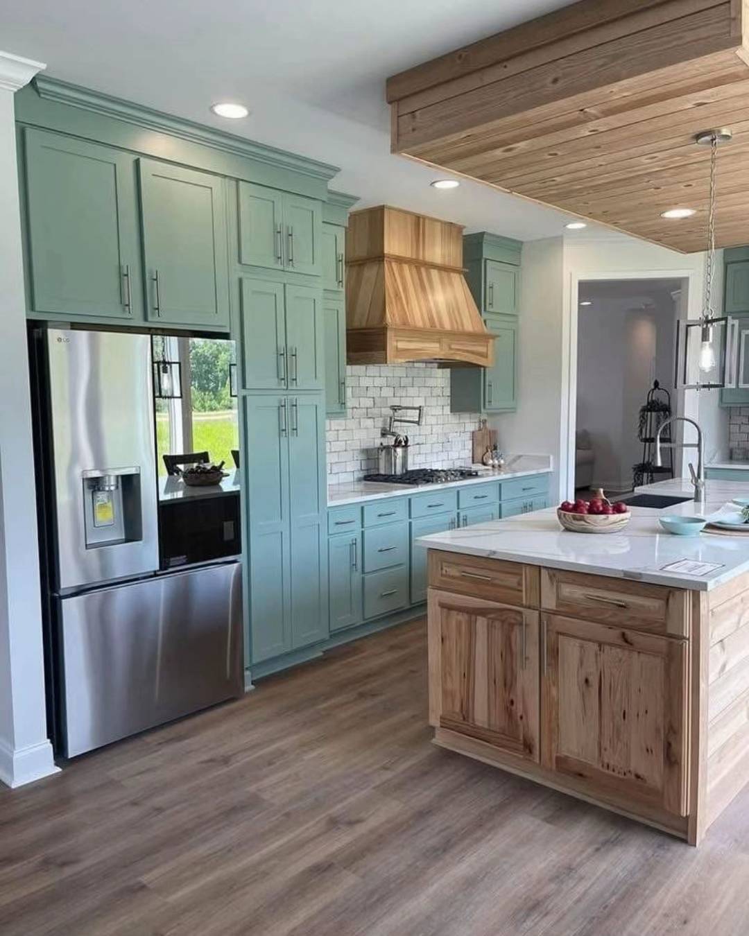 Spacious kitchen with mint green cabinets, a wooden range hood, and stainless steel fridge. Central island with fruit bowl, wood accents, modern feel.
