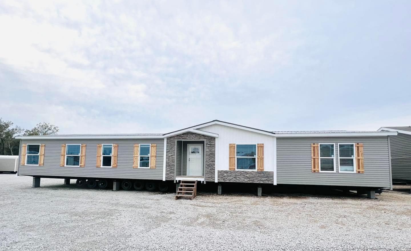 Wide-angle view of a modern manufactured home on a gravel lot. Features gray siding, white trim, and wooden shutters, conveying a neutral, welcoming tone.