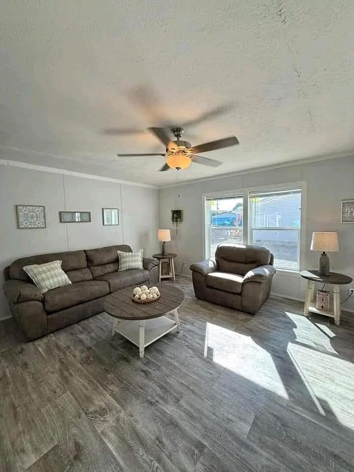 Cozy living room with brown sofas, light gray walls, large windows, and wooden floors. A ceiling fan and lamps add warmth and a tranquil ambiance.