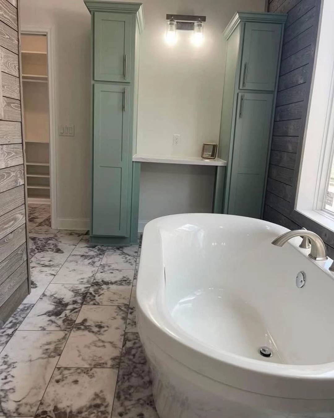 Luxurious bathroom with a white freestanding bathtub on marbled tile floor. Light green cabinets and a modern light fixture create a calm, elegant atmosphere.