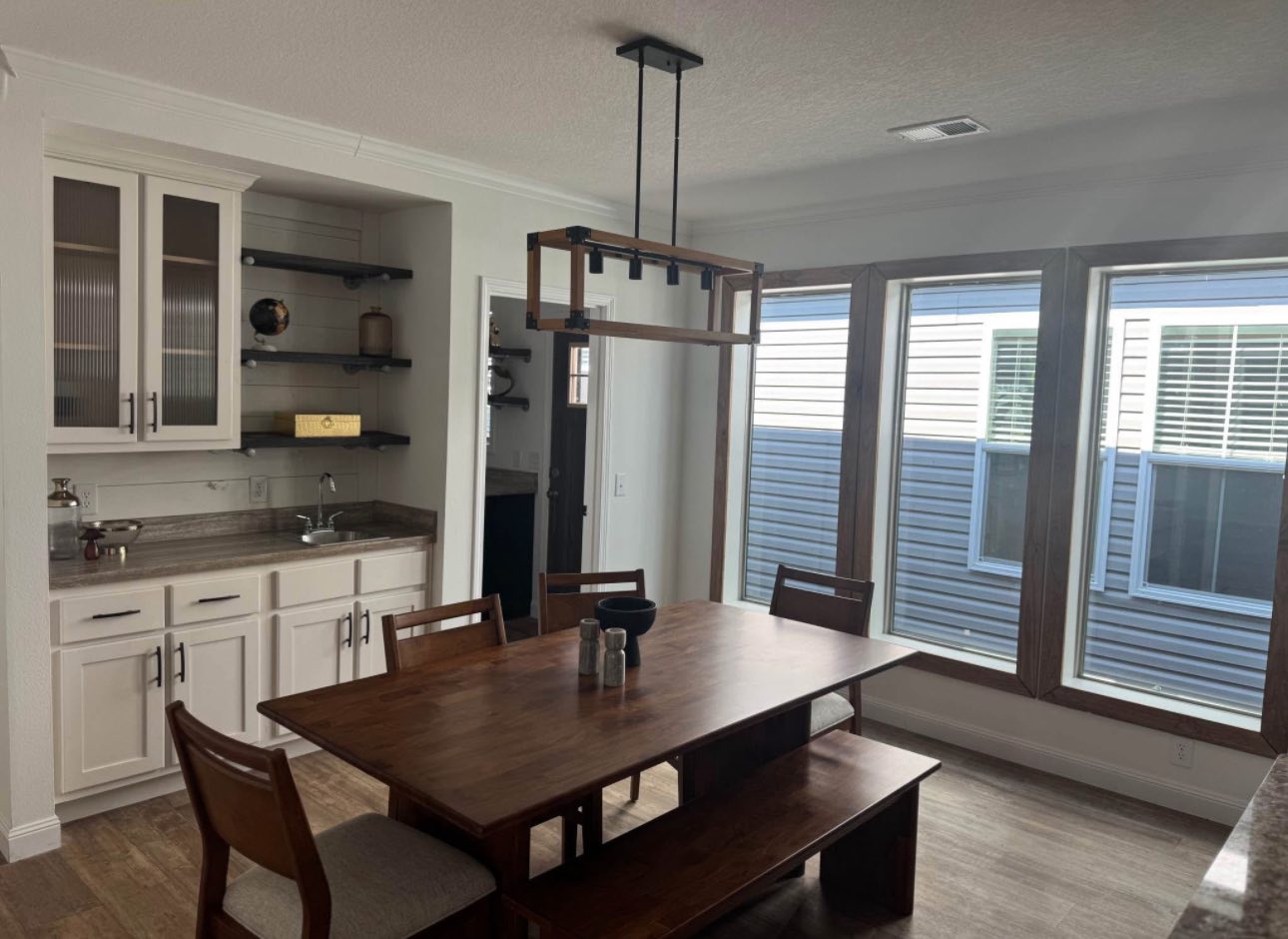 A bright dining room with a rustic wooden table and benches under a modern rectangular chandelier. Large windows let in natural light. A white cabinet and sink are on the left.
