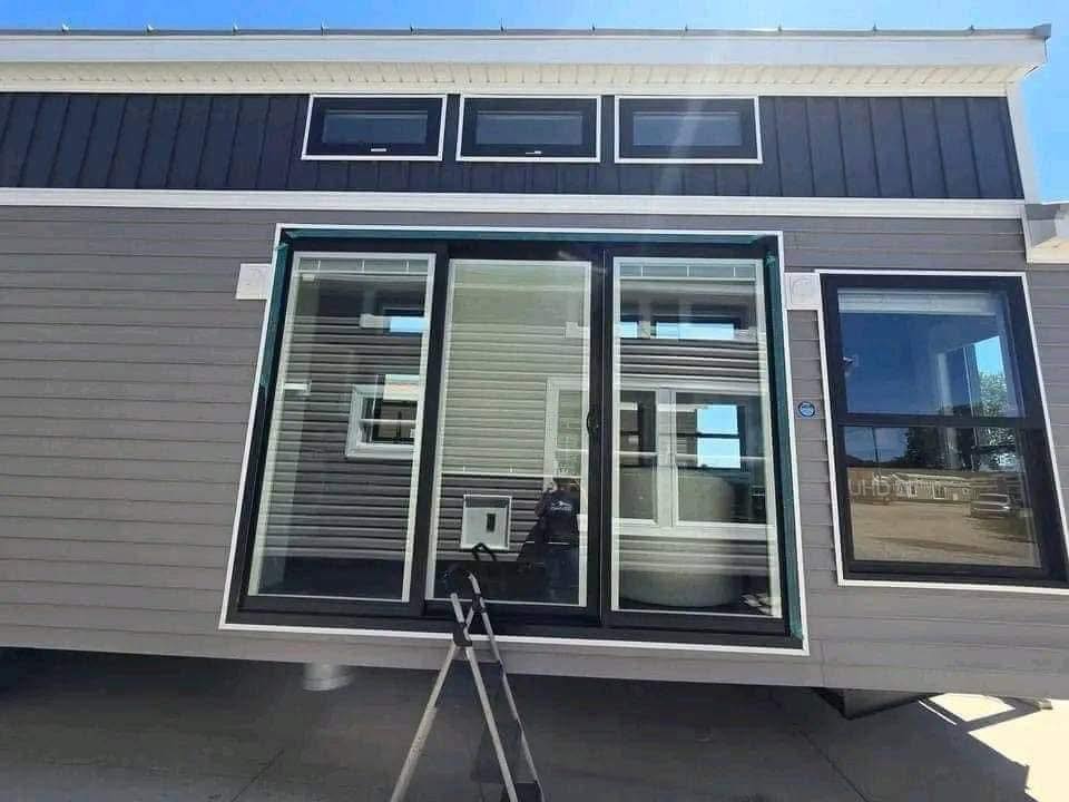 Modern tiny home exterior with large glass sliding doors and windows reflecting sunlight. A small ladder leans against the house. Sunny day.