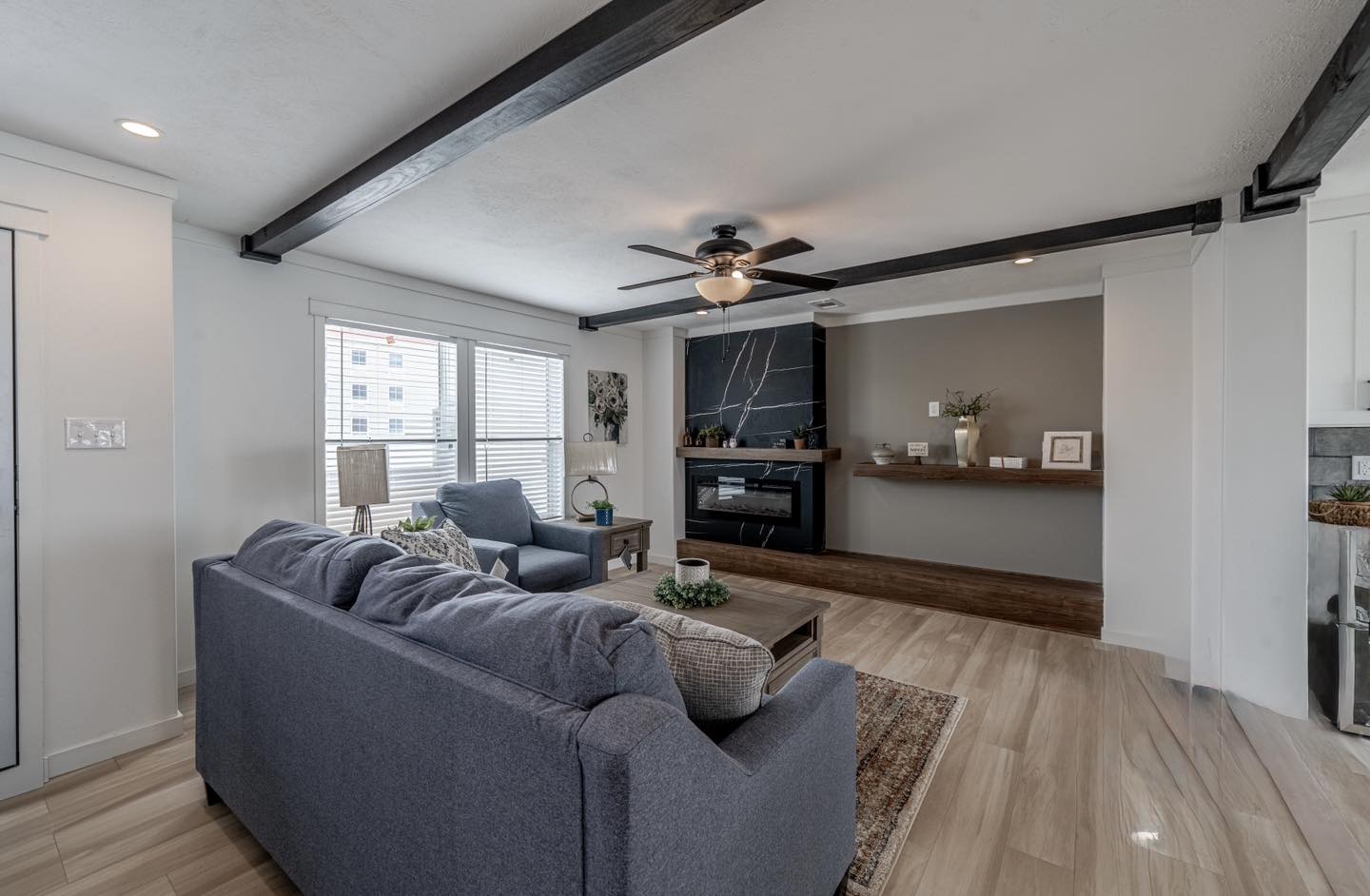 Modern living room with light wood floors, dark ceiling beams, blue sofas, and a black fireplace. Large window brightens the cozy space.