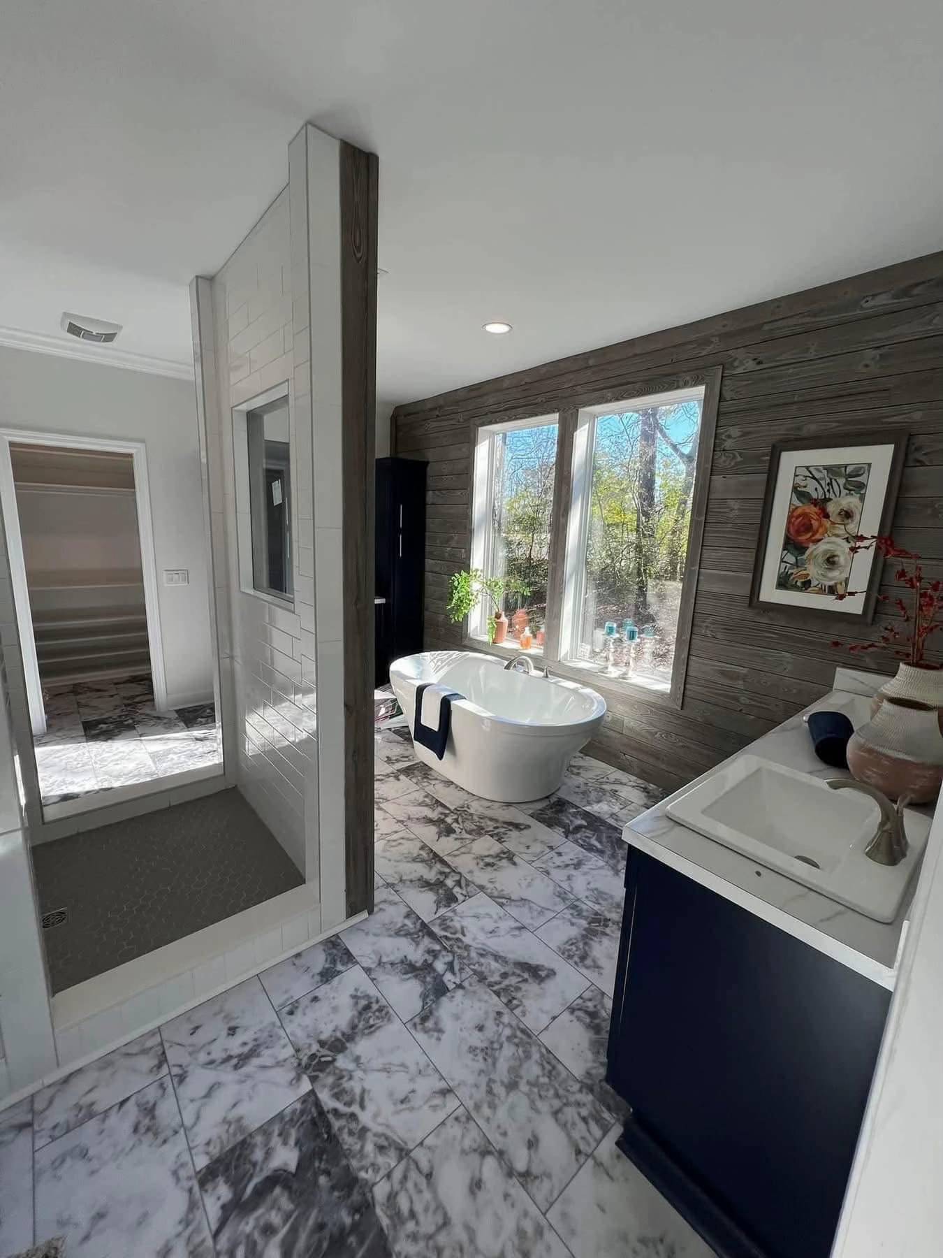 Modern bathroom with marble tile, featuring a freestanding tub under large windows, wood-paneled walls, a walk-in shower, and a dark vanity.