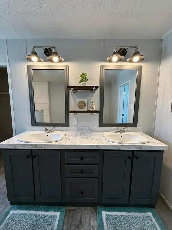 A modern bathroom with a double sink vanity. Two mirrors have overhead lights, and a central shelf holds a plant and decor. Dark cabinetry contrasts with the light walls, and two teal rugs add color.