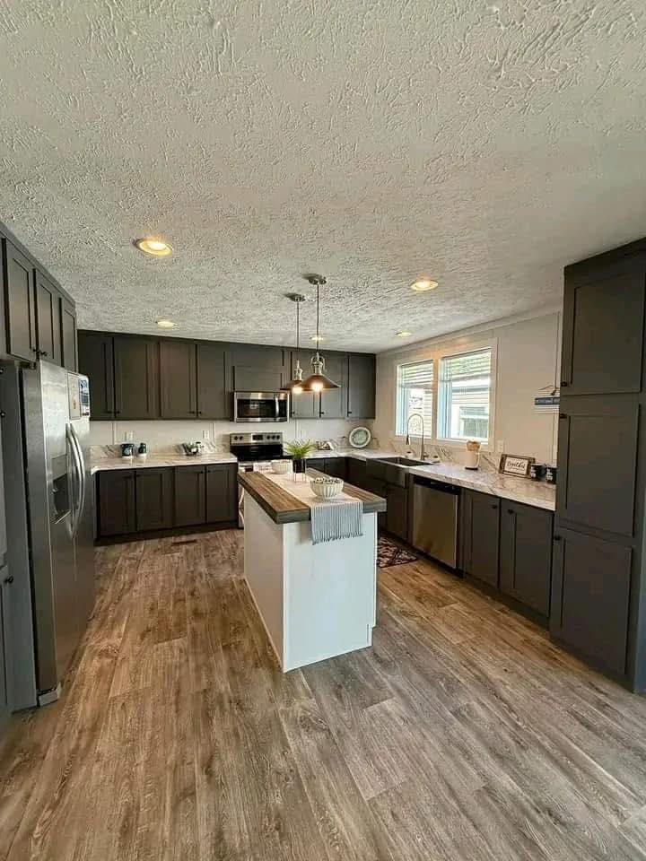 Spacious kitchen with dark cabinets, wood flooring, and a white island under pendant lights. Natural light from a window and stainless steel appliances.