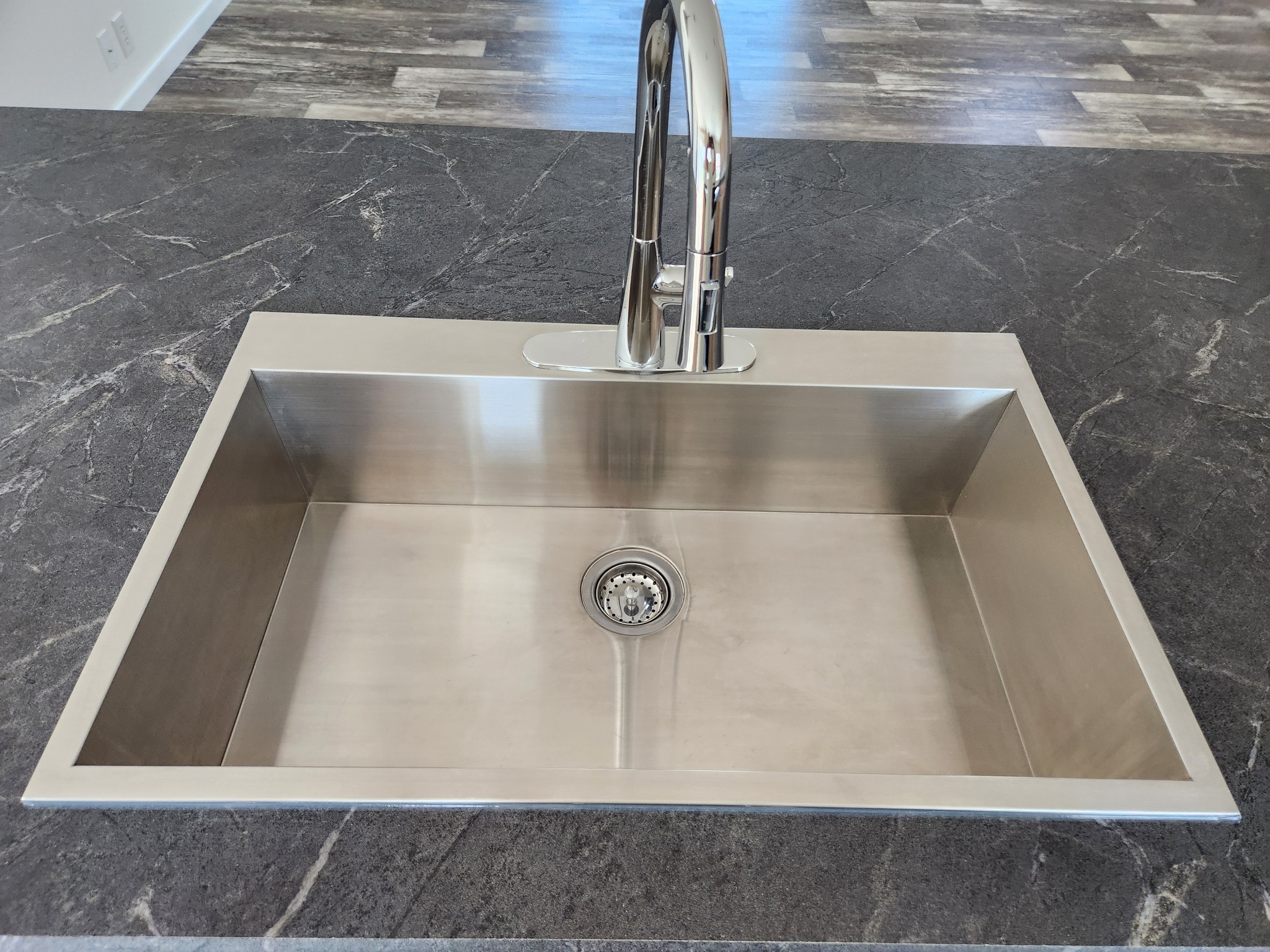 Stainless steel kitchen sink with a modern chrome faucet set in a dark marble countertop. Wood flooring is partially visible in the background.