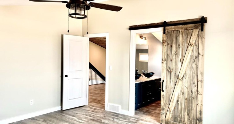 A modern room features light wood flooring, a ceiling fan, and beige walls. An open white door leads to a hallway, and a rustic barn door reveals a bathroom.