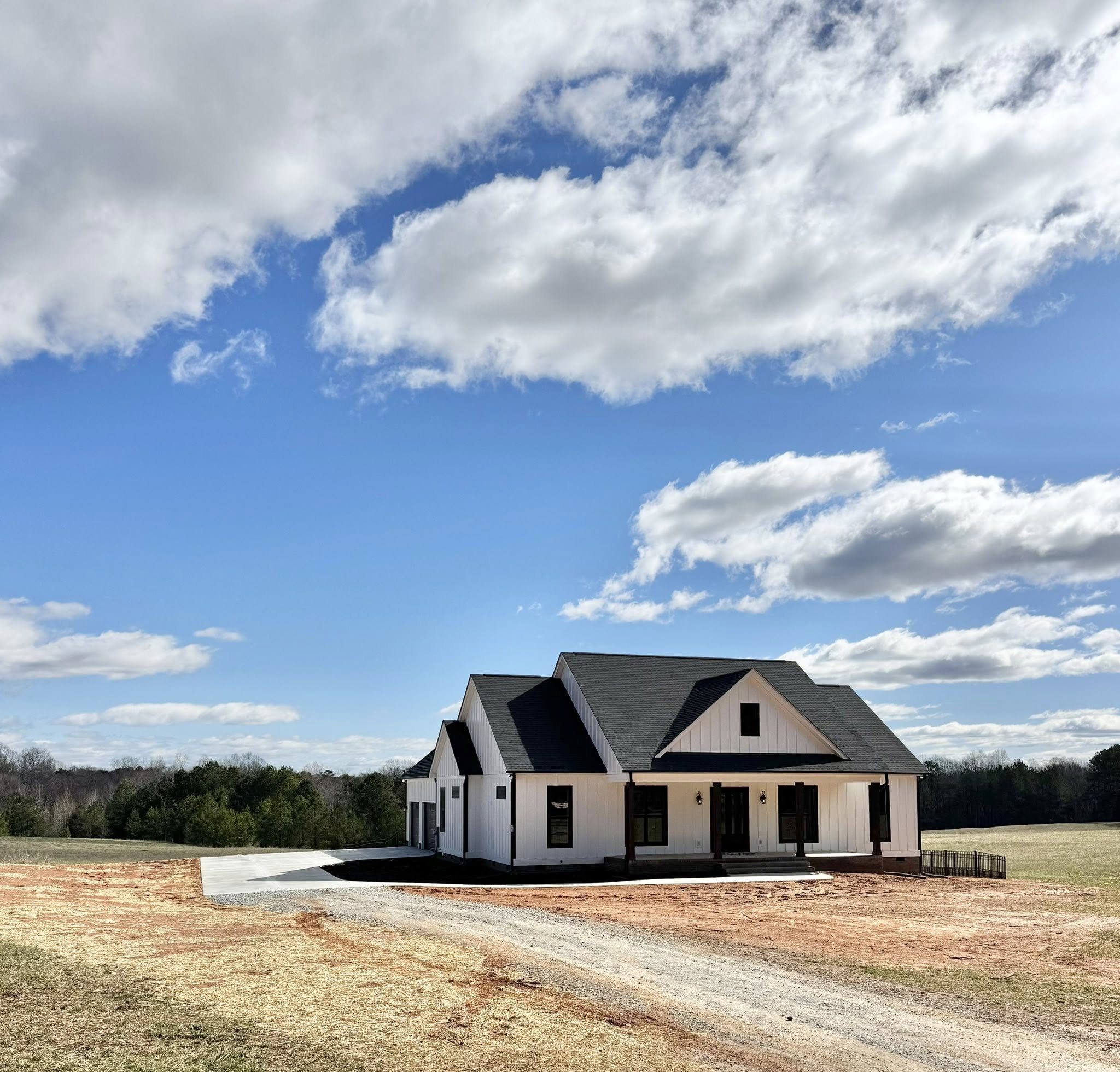 A white farmhouse with a dark roof sits in an open field under a partly cloudy blue sky. The setting is peaceful and rural, with trees in the distance.