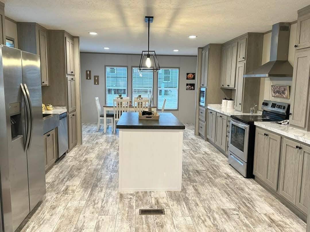 Modern kitchen with light wood cabinets, stainless steel appliances, central island with dark countertop, and wood-textured tile flooring. Bright with natural light from large windows. Cozy dining area in the background.